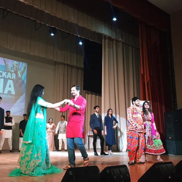 A couple dressed in vibrant traditional attire performs a dance on stage, with the woman wearing a green embroidered dress and the man in a pink kurta. Other performers and audience members are visible in the background, set against a curtain and a screen displaying text.