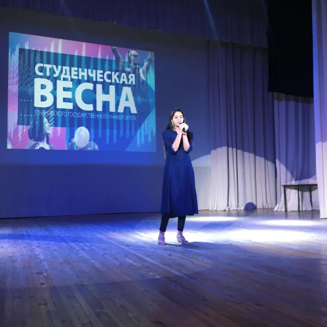 A person in a blue dress stands on a stage, holding a microphone and performing or speaking. The background features a large screen displaying the text "Студенческая Весна" (Student Spring) along with colorful graphics. The setting appears to be a formal event or concert in an auditorium.