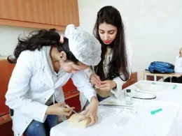 Two individuals in white lab coats working at a table, with one person demonstrating or teaching the other how to use medical equipment. The setting appears to be an educational or clinical environment.