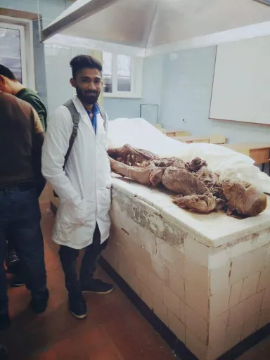 A person in a white lab coat stands next to a table with a preserved anatomical specimen, likely used for medical or scientific study. The setting appears to be a laboratory or educational environment.