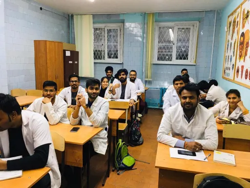 A group of students in white lab coats sitting at desks in a classroom, with one student taking a selfie. The setting includes wooden desks, chairs, and educational materials, indicating a medical or scientific learning environment.