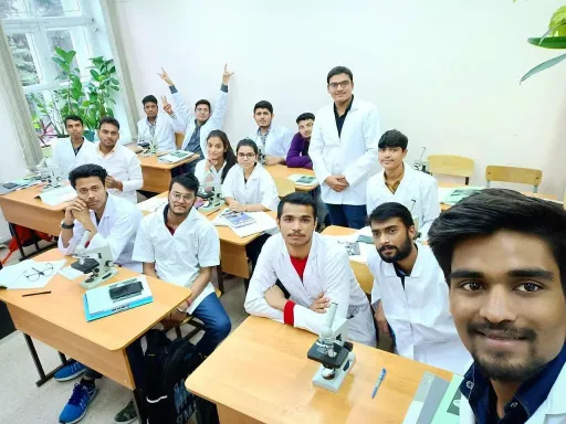 A group of students in white lab coats sitting at desks in a classroom, with one student taking a selfie. The setting includes microscopes and books on the tables, indicating a medical or scientific learning environment.