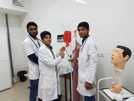 Three individuals in white lab coats standing in a clinical setting, with one person demonstrating an IV setup using a blood bag. A mannequin head is visible on the right, and medical equipment is present in the background.