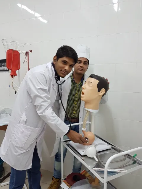 A medical student in a white lab coat using a stethoscope on a mannequin head, with another individual standing behind. The setting appears to be a clinical or training environment, featuring medical equipment and supplies.