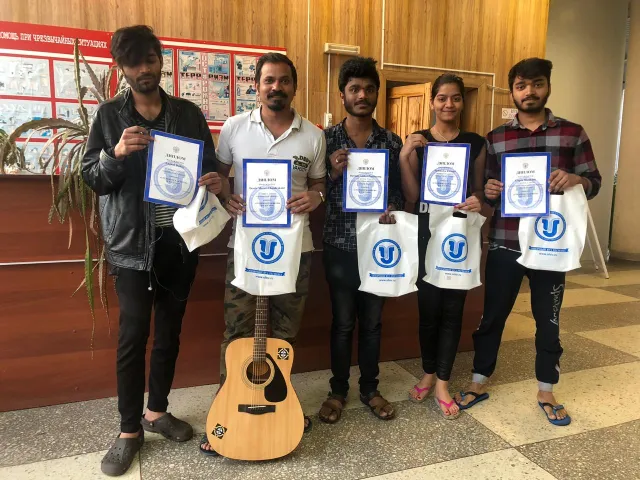 A group of five individuals standing indoors, holding certificates and branded bags. One person is holding an acoustic guitar, and the background features a bulletin board with posters.