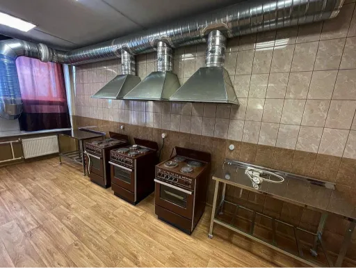A commercial kitchen with multiple stoves, a stainless steel range hood, tiled walls, and wooden flooring. The space includes a countertop and visible ventilation pipes.
