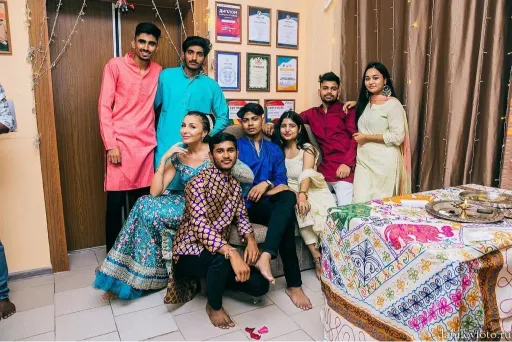 A group of people dressed in traditional attire posing together indoors, with decorative elements such as a patterned tablecloth and framed certificates on the wall.