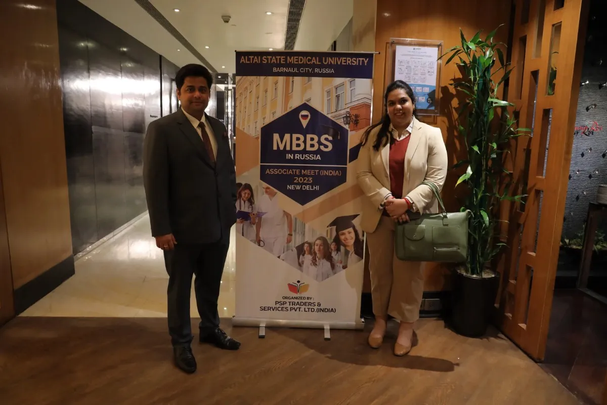 Two individuals stand in front of a banner for Altai State Medical University's MBBS program in Russia, featuring an "Associate Meet (India) 2023" in New Delhi.