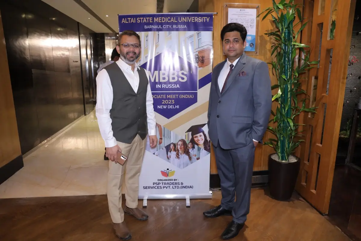 Two individuals are standing in front of a promotional banner for Altai State Medical University, Barnaul City, Russia. The banner highlights the "MBBS IN RUSSIA" program and mentions an "Associate Meet (India) 2023" held in New Delhi. The event is organized by PSP Traders & Services Pvt. Ltd. (India). The setting appears to be indoors with modern decor, including wooden flooring and a potted plant nearby.