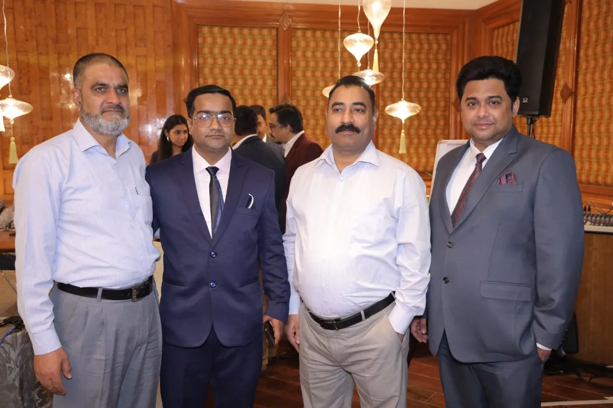Four individuals are standing together in a formal setting, dressed in business attire. The background features warm wooden paneling and decorative lighting, suggesting an elegant indoor event or meeting space