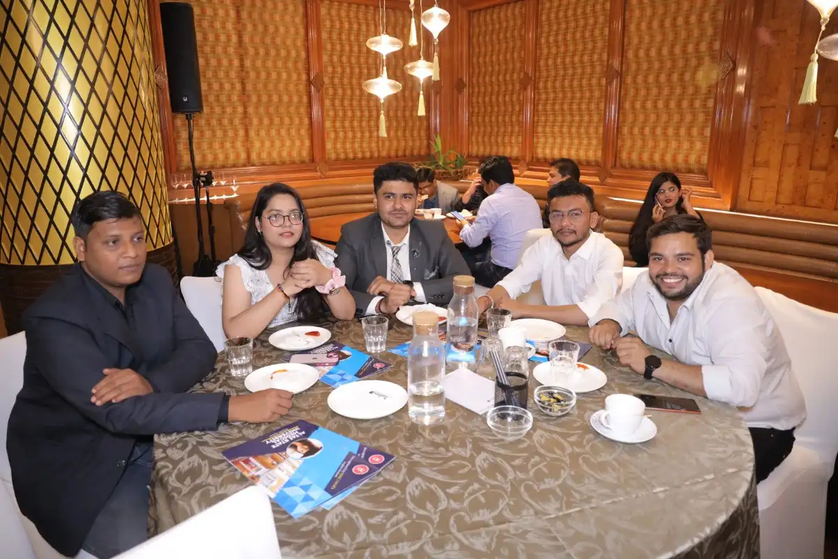 A group of individuals is seated around a round table in an elegant indoor setting, likely during a formal event or meeting. The table is set with plates, glasses, water bottles, and brochures, indicating a professional gathering. The background features warm wooden paneling and decorative lighting, adding to the sophisticated ambiance.