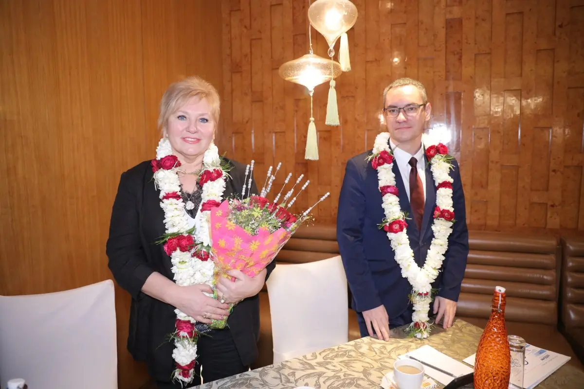 A man and a woman are seated at a table in an elegant indoor setting, both adorned with traditional flower garlands. The woman holds a bouquet of flowers wrapped in pink paper, while the table is set with cups, papers, and decorative items. The background features warm wooden paneling and hanging lanterns, creating a formal and welcoming atmosphere