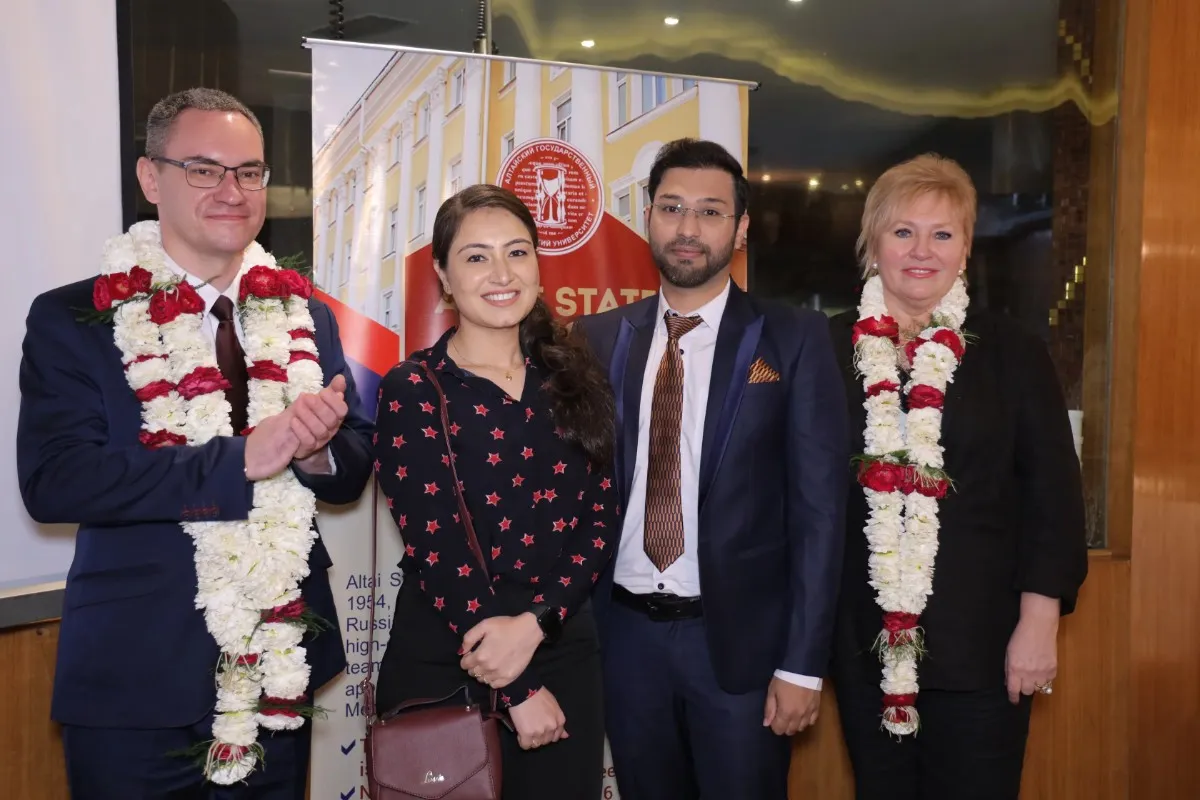 Four individuals are standing together at a formal event, all wearing traditional flower garlands. They are dressed in business attire, with one person holding a handbag. The background features a banner promoting Altai State Medical University, indicating an educational or professional gathering.