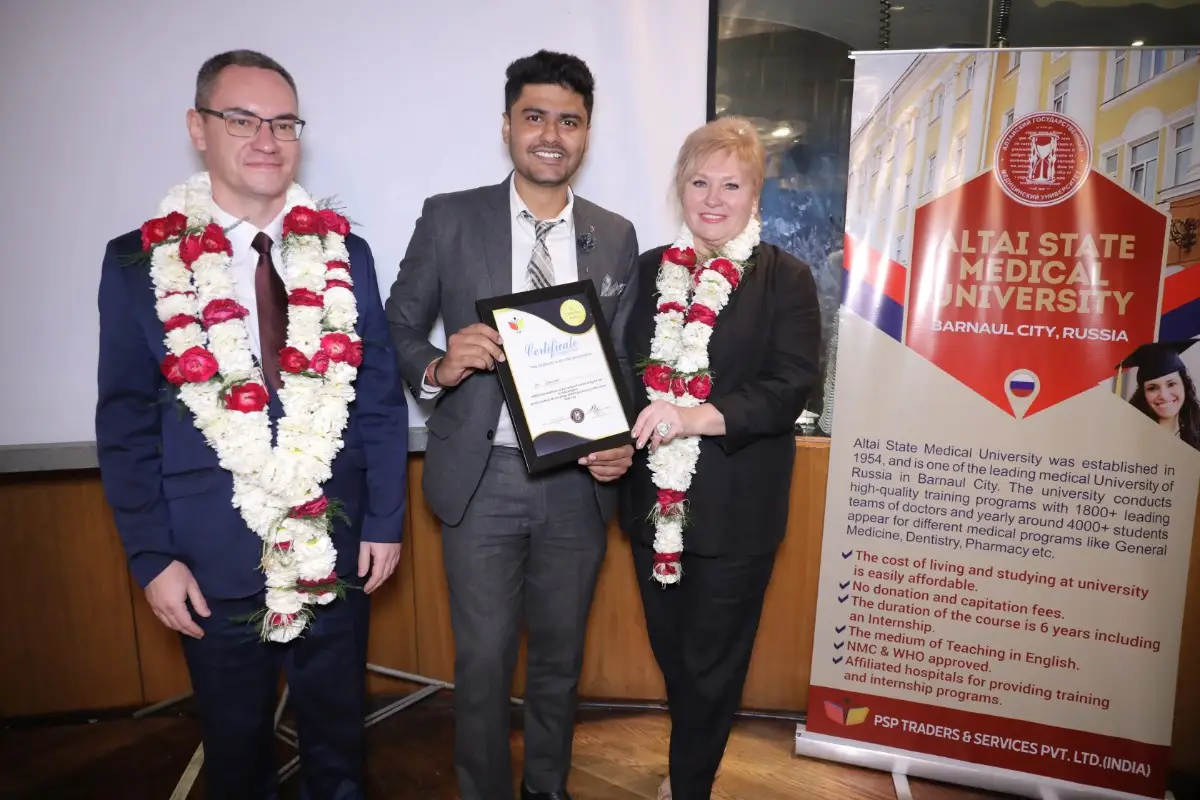 Three individuals, adorned with flower garlands, stand together at an event. The central person holds a certificate, while the backdrop features a banner for Altai State Medical University in Barnaul City, Russia.