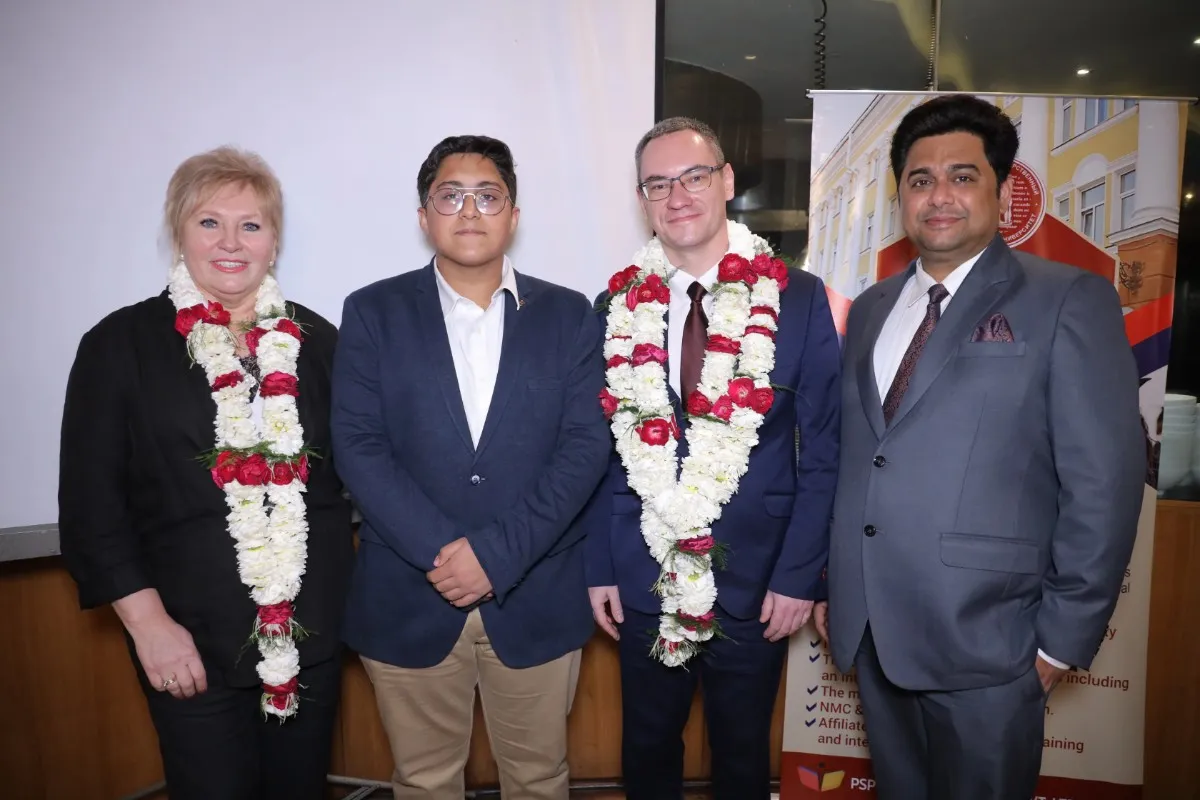 Four individuals, dressed in formal attire and wearing flower garlands, stand together at an event. The background includes a banner with text related to Altai State Medical University.