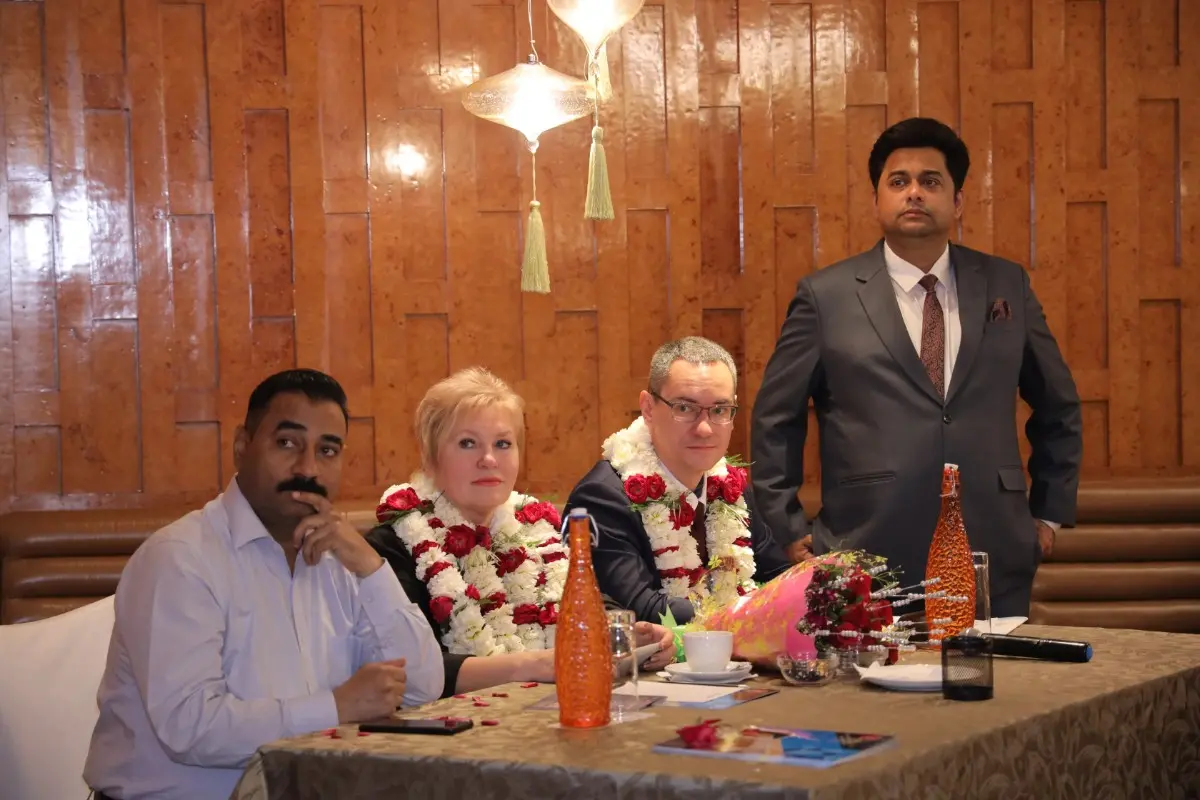 A group of individuals, some wearing flower garlands, are seated at a table in a warmly lit room with wooden paneling. The setting suggests a formal or celebratory event, with decorative elements like a hanging light and floral arrangements.