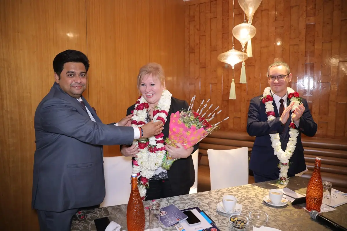 A formal event where a man presents a bouquet of flowers to a woman wearing a flower garland, while another man in the background claps. The setting features warm lighting, wooden paneling, and a table with decorative items.